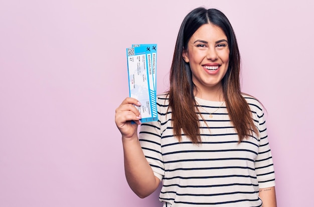 Young beautiful tourist woman holding airline boarding pass over isolated pink background looking positive and happy standing and smiling with a confident smile showing teeth