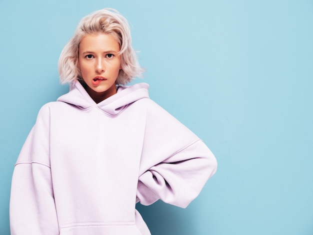 Young beautiful upset female in trendy summer hoodie Sexy carefree woman posing near blue wall in studio Sad blond model having fun and going crazy Cheerful and happy biting her lips