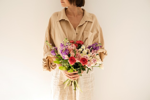 Young beautiful woman in beige muslin shirt hold bouquet of colourful roses flowers against white wall Beautiful holiday celebration floral composition