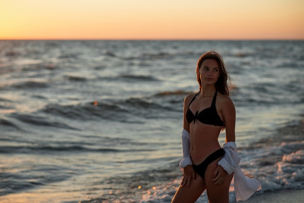 Young beautiful woman in black bikini and white shirt on a tanned body on the beach at sunset. Soft selective focus.