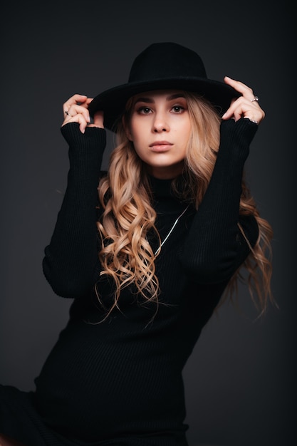 Young beautiful woman in black hat sitting on black wall