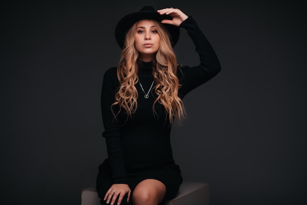 Young beautiful woman in black hat sitting on black wall