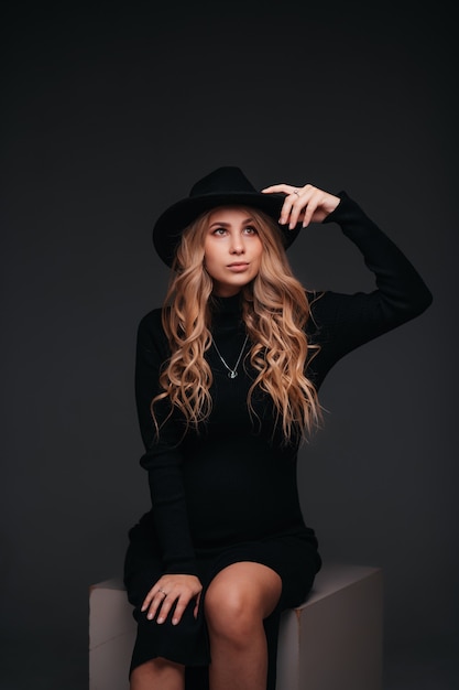 Young beautiful woman in black hat sitting on black wall