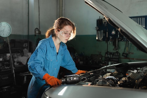 Young beautiful woman car mechanic is standing near the open hood of a car and makes a technical inspection at a service station