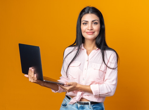 Young beautiful woman in casual clothes holding laptop happy and positive looking at front smiling confident standing over orange wall