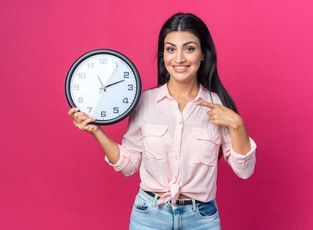 Young beautiful woman in casual clothes holding wall clock pointing with index finger at it smiling cheerfully standing on pink