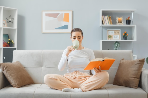 Young beautiful woman in casual clothing sitting on a couch at home interior drinking tea and reading a book looking confident spending weekend at home