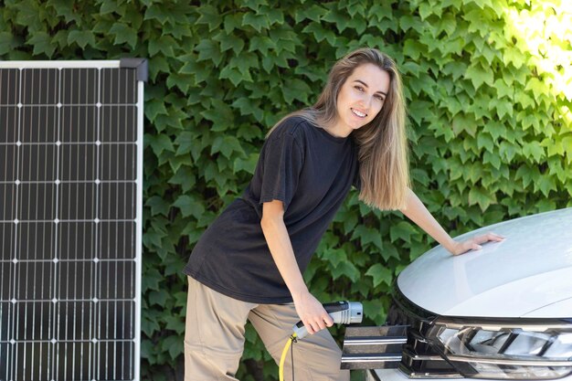 Young beautiful woman charging parked electric car from solar power station with photovoltaic panel