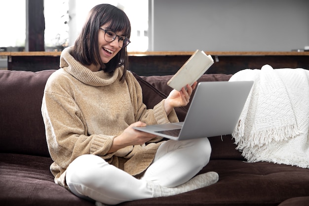 Young beautiful woman in a cozy sweater with a book in her hands and a laptop on the couch at home. Online learning