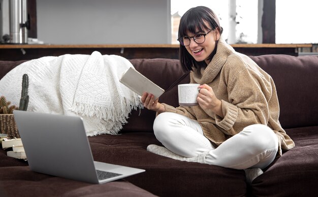 Young beautiful woman in a cozy sweater with a cup and a book in her hands and a laptop on the couch at home. Online learning concepts.