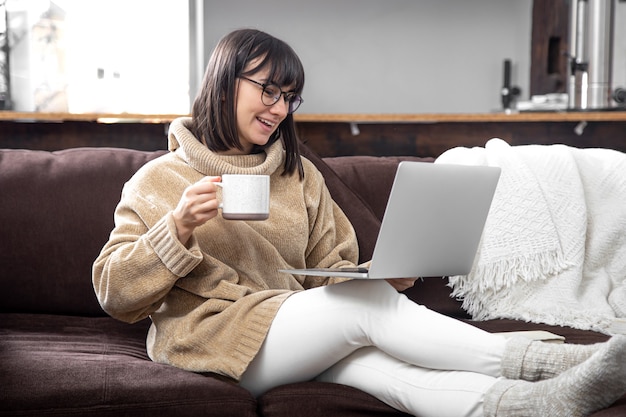 Young beautiful woman in a cozy sweater with a mug of drink works at a laptop. Remote work at home and freelancing.