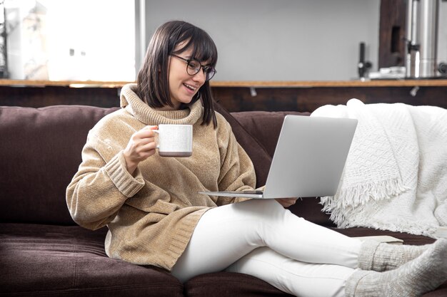 Young beautiful woman in a cozy sweater with a mug of drink works at a laptop. Remote work at home and freelancing.