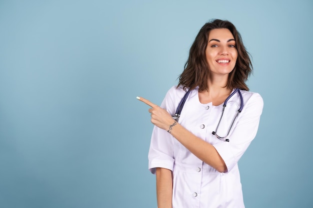 Young beautiful woman doctor in a lab coat on a blue background smiles, and points her finger to the left on an empty space