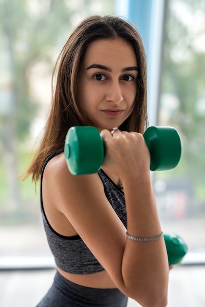 Young beautiful woman doing exercise with dumbbells while standing in front of window at gym