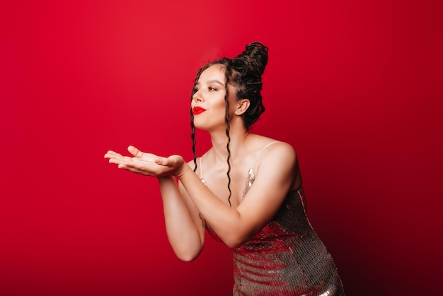 A young beautiful woman in a dress with dreadlocks sends an air kiss on a red background advertising photo space for text