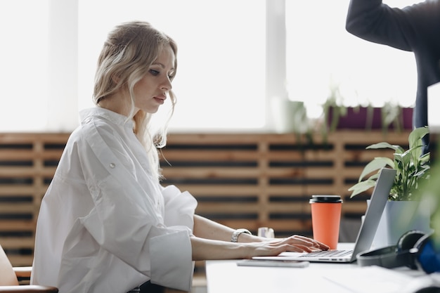 Young beautiful woman dressed in white shirt is working at the laptop sitting at the desk in a light modern open space office