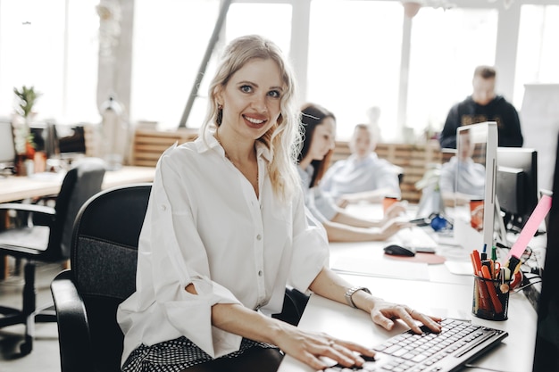 Young beautiful woman dressed in white shirt is working at the laptop sitting at the desk in a light modern open space office