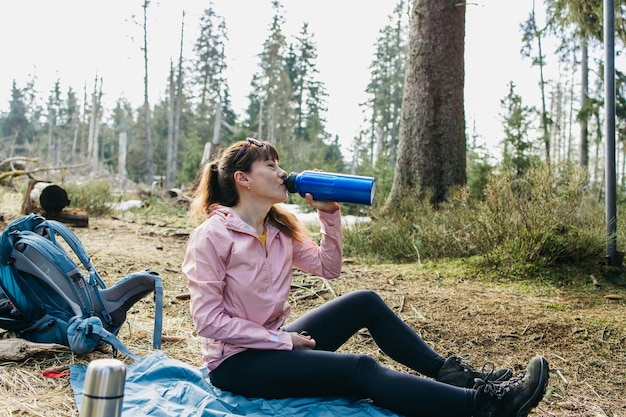 Photo young beautiful woman drinking hot tea or coffee from a thermos in the forest on a walk hiking