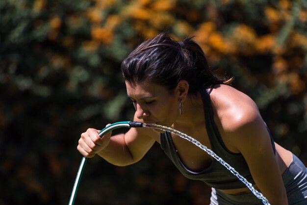 Young and beautiful woman drinking water from the hose after exercising thirsty sunny hot day