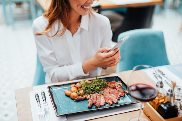 Young beautiful woman enjoying in tasty and nicely decorated meal. She is sitting in expensive restaurant and using her smart phone for food photography.