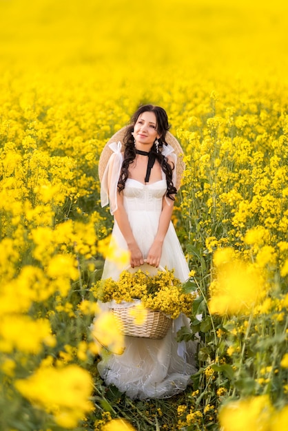 Young beautiful woman in a field with a large basket of yellow flowers A girl in a long white dress collects rapeseed