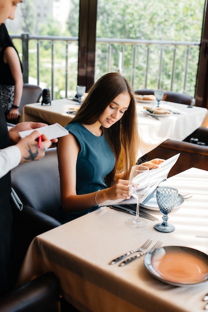 A young beautiful woman in a fine restaurant looks at the menu and makes an order to a young waiter in a stylish apron. Customer service.