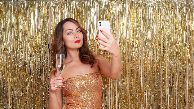 Young beautiful woman in a golden dress drinks champagne, makes a selfie on a smartphone, having fun at a party on a shiny gold background