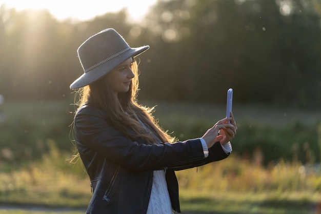 A young, beautiful woman in a hat smiles, takes a selfie and takes a video in the park on her smartphone in the bright radiance of the rays of the sunset rays