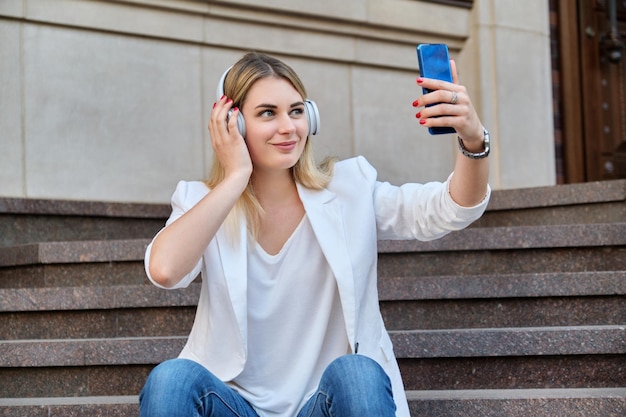 Young beautiful woman in headphones with smartphone resting sitting on steps in city