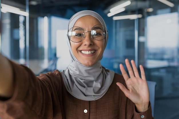 Young beautiful woman in hijab looking at smartphone camera talking with friends on video call