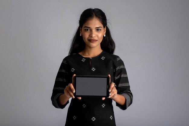 Young beautiful woman holding and showing blank screen of smartphone or mobile or tablet phone on a gray.