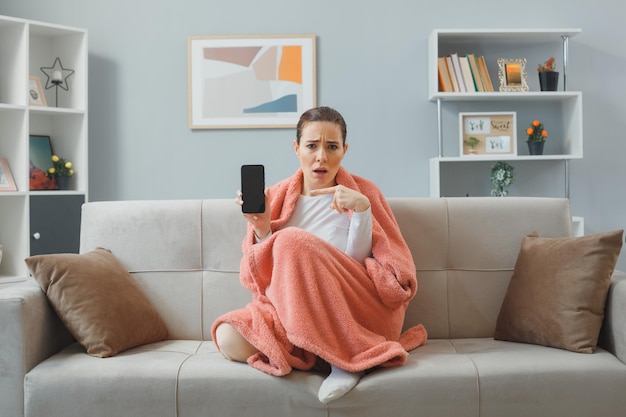 young beautiful woman in home clothes sitting on a couch under blanket at home interior using smartphone showing at camera pointing with index finger at it looking confused spending time at home