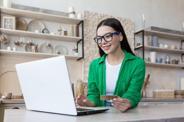 Young beautiful woman at home in the kitchen in glasses and a green shirt makes online purchases in