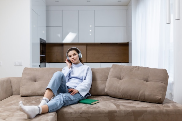 Young beautiful woman is resting at home, sitting on the couch, and listening to music on headphones