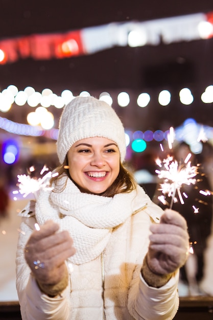 Young beautiful woman in knitted hat and scarf standing in the city with bengal light, sparkler. Concept celebration and christmas.