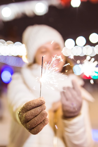 Young beautiful woman in knitted hat and scarf standing in the city with bengal light sparkler