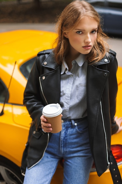 Young beautiful woman in leather jacket leaning on yellow sport car holding cup of coffee to go in hand while thoughtfully 