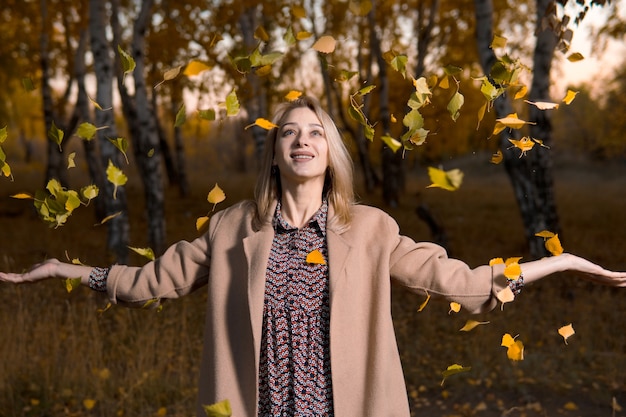 Young beautiful woman in long coat throwing leaves in autumn forest.