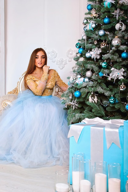 Young and beautiful woman in long dress with Christmas gift in her hands sitting near Christmas tree