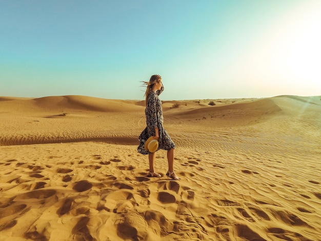 Young beautiful woman in long dress and with a hat in the middle of the Dubai desert with sunlight