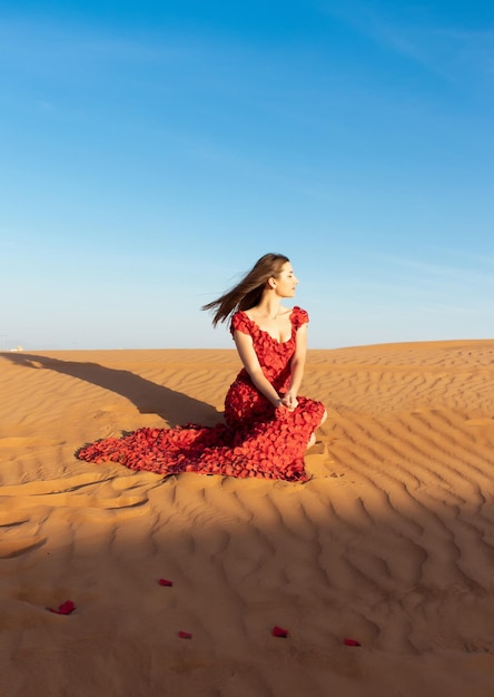 Young beautiful woman in long red dress with red rose petals among the desert Desert rose conception