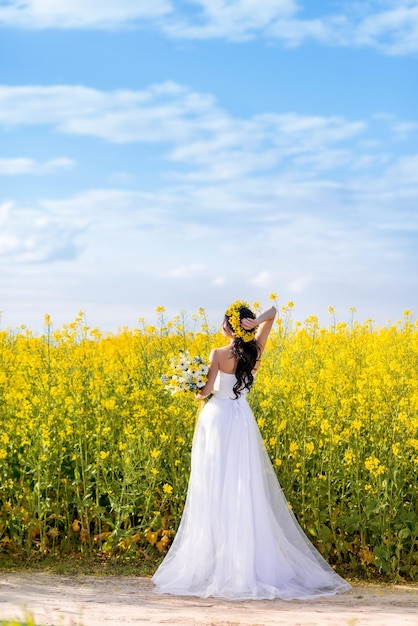 Young beautiful woman in a long white dress is standing on a rural country road A bride with a bouquet in a yellow flowering rapeseed field Against a beautiful blue sky