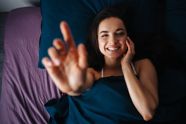 Young beautiful woman in morning bed at home. Positive cheerful woman smile. Touch her face and show blurred hand. Picture concentrated on woman.