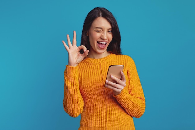 Young beautiful woman in orange sweater showing ok gesture while using