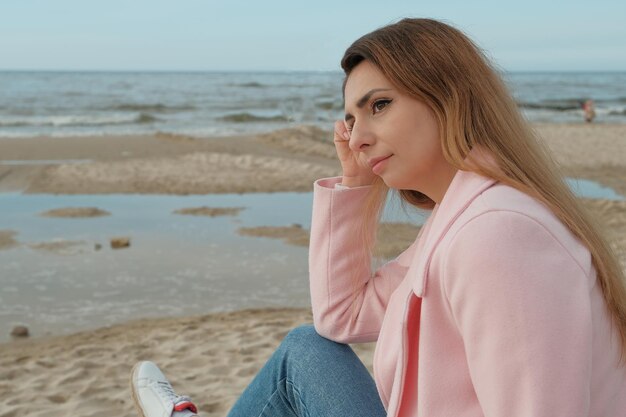 A young beautiful woman in a pink coat stands on the sea or ocean shore with a cup of coffee and enj