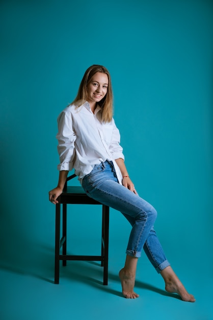 Young beautiful woman posing on a chair in a white shirt and jeans on a blue wall barefoot