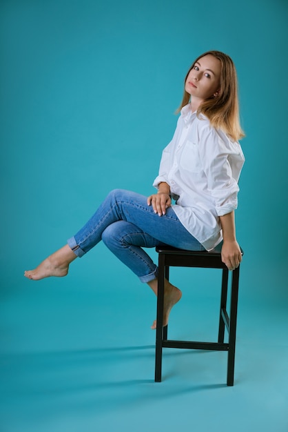 Young beautiful woman posing on a chair in a white shirt and jeans on a blue wall barefoot