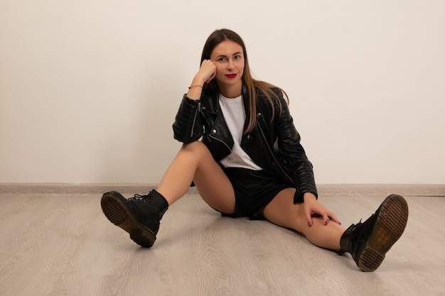 Young beautiful woman posing sits in studio in black leather shorts, boots and jacket