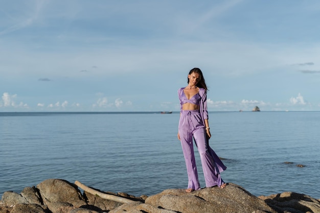 Young beautiful woman in a romantic mood lilac silk clothes on the beach against the backdrop of the sea and stones at sunset