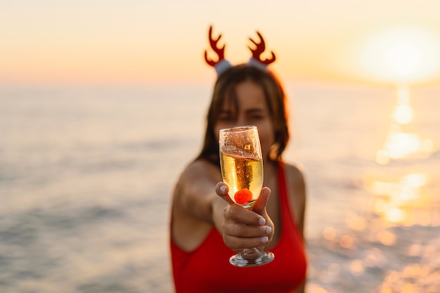 Young beautiful woman in a santa hat with a glass of champagne in her hands walk on the beach